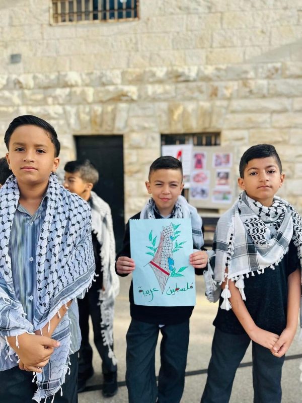 ثلاثة طلاب بالكوفية، أحدهم يحمل رسمة، في ساحة المدرسة