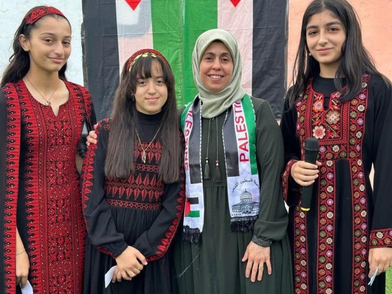 ثلاث طالبات بأثواب مطرّزة تقليدية يقفن مع معلمة ترتدي وشاح فلسطين