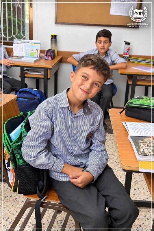 A smiling student sits at his desk with his school bag