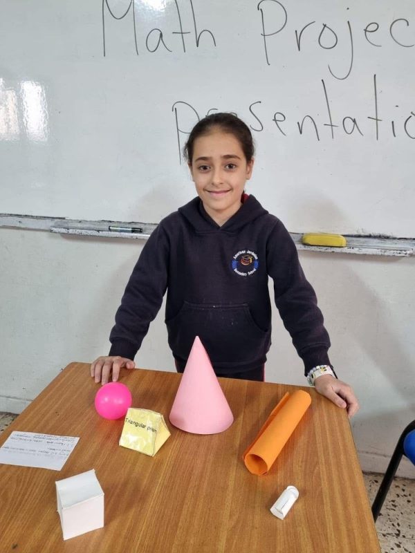 A student presents 3D shapes she made, under the heading “Math Project Presentation”